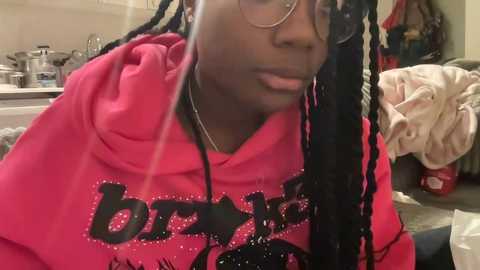 Media: Video of a young Black person with dreadlocks, wearing glasses and a pink hoodie with \"Bread\" printed in bold black letters, sitting in a cluttered kitchen.