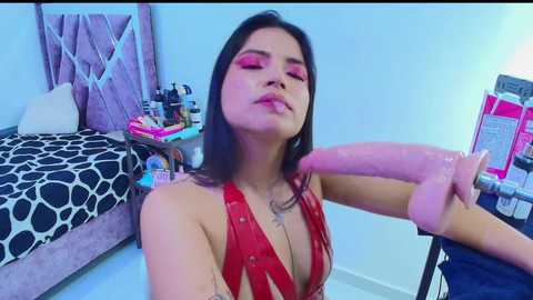 Media: A video of a young woman with long dark hair and fair skin, wearing a red leather halter top, is seen in a bedroom holding a large, erect, pink dildo.