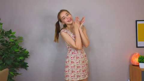 Media: Video of a smiling young woman with light skin and long brown hair in a floral dress, standing against a beige wall with a potted plant and a lamp in the background.