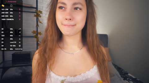 Media: Video of a young woman with long, wavy brown hair, fair skin, and a subtle smile, wearing a white lace camisole. Background shows a dimly lit bedroom with a bed and a plant.