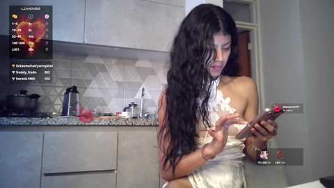 Media: Video of a woman with long black hair, wearing a white lace dress, checking her smartphone in a modern kitchen with gray cabinets and a tiled backsplash.