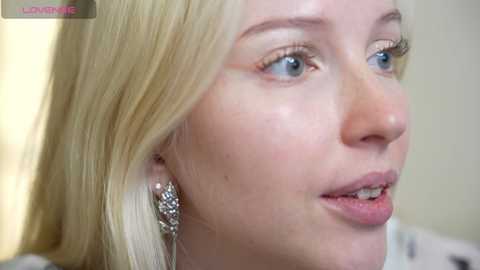 Media: Close-up video of a fair-skinned, blonde woman with blue eyes and a small mole on her cheek, wearing a sparkling diamond earring. Background is blurred, focusing on her facial features.