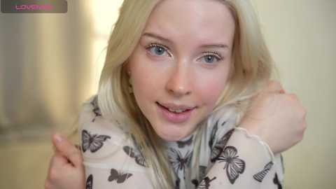 Video of a young, fair-skinned woman with long, straight blonde hair, blue eyes, and a slightly open mouth, wearing a white blouse with butterfly patterns. She's indoors, with a blurred, beige background.