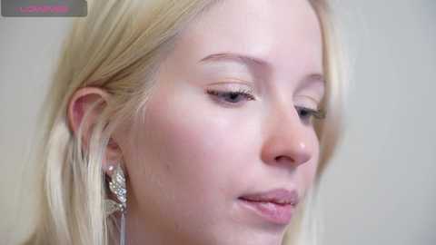 Video of a fair-skinned, blonde woman with straight hair, wearing silver earrings, looking down. The background is a plain, light-colored wall.