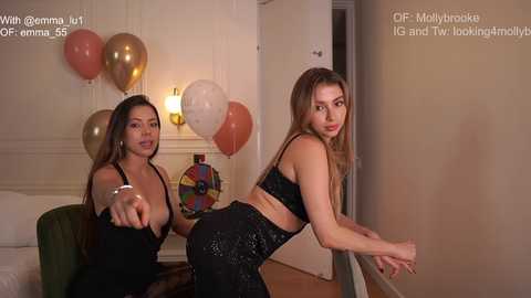 Media: A video of two women, one in a black sequined dress, the other in a black top and pants, posing in a modern living room with a white door and a colorful dartboard.