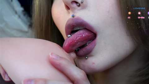 Media: Video of a close-up of a fair-skinned woman with a pierced tongue and multiple facial piercings, licking the nipple of a person, likely male, with a visible penis.