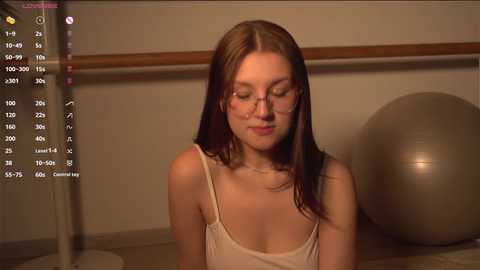 Media: A video of a fair-skinned, young woman with long brown hair, wearing a white tank top, sitting on a bed, eyes closed, serene expression, in a dimly lit room with a large, silver exercise ball in the background.