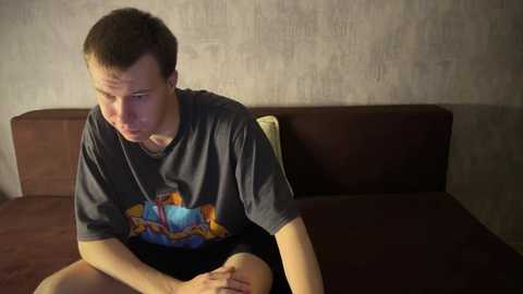 Media: A video of a young man with short brown hair, wearing a gray T-shirt with a colorful graphic, sitting on a dark brown couch in a dimly lit room with textured gray walls.