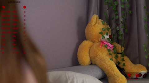 Media: Video of a large, fluffy, golden teddy bear with green vines and pink ribbon on a grey bed, partially blurred figure in the foreground.