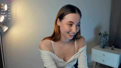 Media: Video of a young woman with light skin, long brown hair, and a white off-shoulder top, smiling in a dimly lit room with a white dresser and plant.