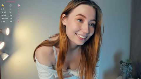 Media: Video of a young Caucasian woman with long, straight, light brown hair and fair skin, smiling widely. She wears a white off-shoulder top and sits against a blurred, softly lit background.