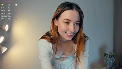 A video of a young woman with light skin and long, straight, reddish-brown hair, wearing a white top. She has a warm smile and is indoors with a blurred background.