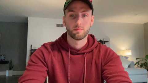 Media: Video of a bearded, fair-skinned man in a red hoodie and cap, standing indoors, with a modern, minimalist living room in the background.