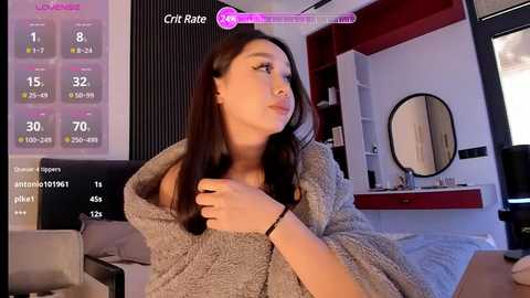 Media: Video of a young woman with long dark hair, wearing a gray robe, in a modern, minimalistic bedroom with a bed, mirror, and digital clock displaying \"Cric Rate.\