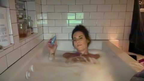 Media: A video of a woman with short dark hair, partially submerged in a bathtub, holding a blue bottle, in a white-tiled bathroom.