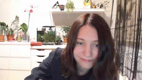 Media: Video of a young woman with wavy brown hair, wearing a black top, sitting in a bright, cluttered kitchen with white cabinets and green plants.