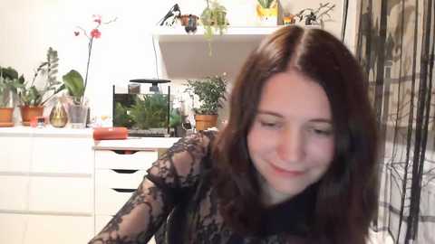 Media: Video of a young woman with long, wavy brown hair, wearing a black lace top, smiling, in a modern, well-lit kitchen with white cabinets, potted plants, and a fish tank.