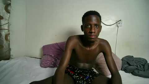 Media: Video of a shirtless young African boy with short hair, sitting on a bed with white sheets, wearing black shorts with red and blue patterns, in a simple room with a white wall and a telephone.