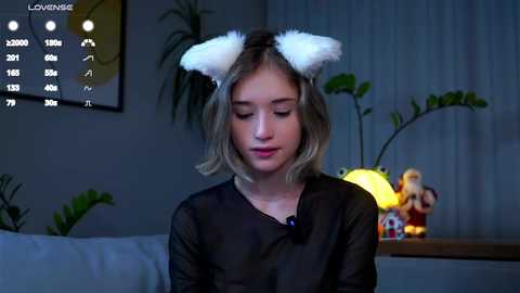 Video of a young woman with blonde hair wearing white cat ears, sitting indoors at a table with a lit candle, plants, and a colorful toy.