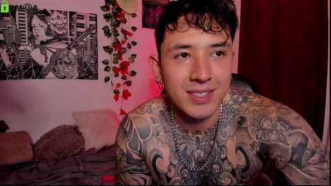 Media: Video of a tattooed, smiling man with short dark hair, wearing a silver chain necklace, in a dimly lit bedroom with black-and-white posters, red rose garland, and a bed with gray sheets.