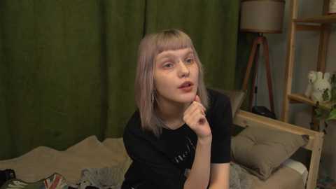 Media: A video of a young, fair-skinned woman with pale pink hair, wearing a black t-shirt, sitting on a bed with green curtains behind her.