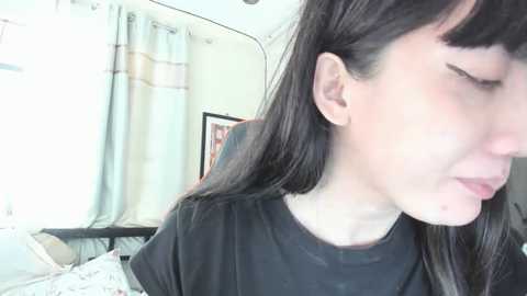 Media: A video of a young woman with long black hair and fair skin, wearing a black t-shirt, looking down with a slight frown, in a bright, softly lit bedroom with light blue curtains.