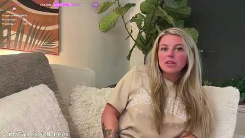 Media: A video shows a blonde woman with long hair, wearing a beige T-shirt, seated on a grey couch with a fluffy white pillow. A green plant and a framed artwork with a palm tree design are in the background.