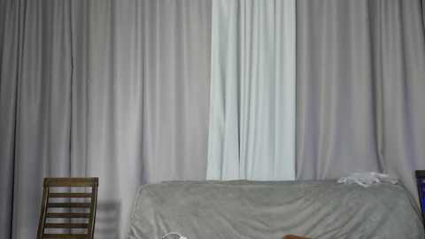 Media: Video of a minimalist room with a single bed covered in a light gray blanket, flanked by two wooden chairs, and a white curtain partially drawn to reveal a window.