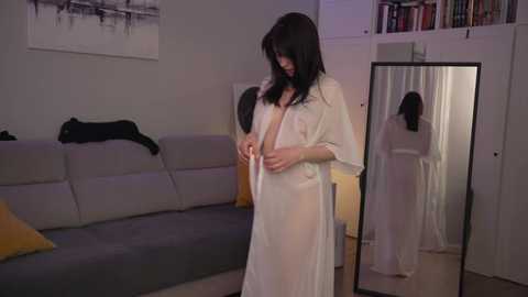 Media: Video of a slender, fair-skinned woman with long black hair in a sheer white robe, standing in a modern living room with a grey couch, a black cat, and a tall mirror.