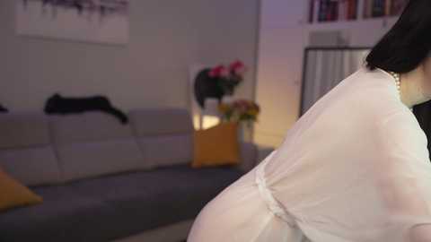 Media: Video of a woman in a white dress sitting on a gray couch, with a black cat on the left and a vase of flowers on the right, in a dimly lit living room.