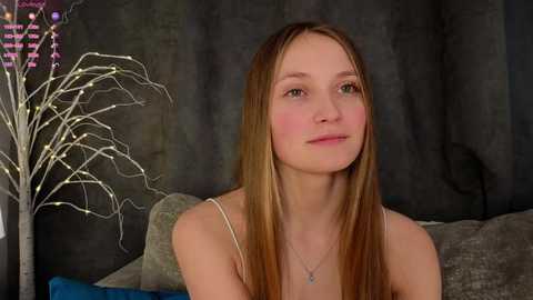 Media: Video of a young Caucasian woman with fair skin and long, straight, light brown hair, wearing a white spaghetti-strap top, sitting against a dark, textured backdrop.