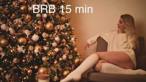 Media: Video of a blonde woman in a white sweater and knee-high socks, seated on a brown couch, gazing at a gold and silver-decorated Christmas tree. Text reads \"BBR 15 min.\