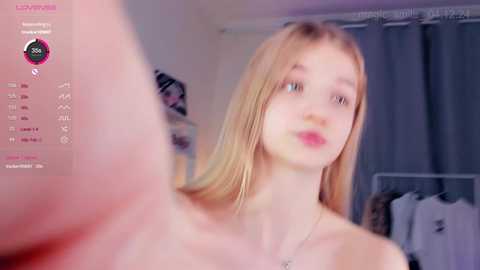 Media: Video of a young, fair-skinned, blonde girl with blue eyes, wearing a strapless top, standing in a bedroom with clothes hanging on a rack.