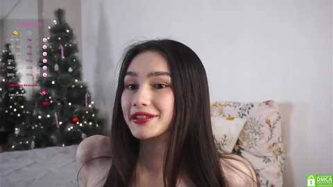 Video of a young woman with long black hair, fair skin, and red lipstick, smiling in a cozy room with a decorated Christmas tree, floral pillows, and a calendar in the background.