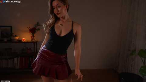 Media: Video of a slender, Caucasian woman with light skin and brown hair, wearing a black tank top and maroon leather mini-skirt, standing in a dimly-lit room with bookshelves and candles.