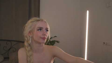 Media: Video of a fair-skinned, blonde-haired woman with a braided hairstyle, wearing a strapless dress, looking upwards at a vertical light source in a dimly-lit room.