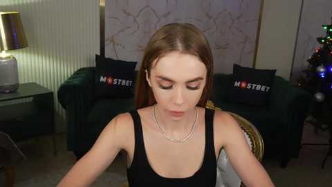 Media: A video of a young woman with fair skin, straight brown hair, wearing a black tank top, seated indoors in a modern living room.