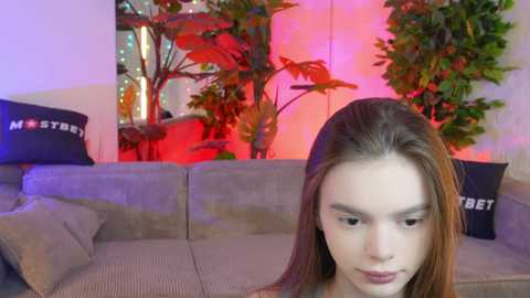 Media: Video of a young woman with long, straight brown hair, light skin, and fair eyebrows, sitting on a beige couch. Background features green plants, a \"HUSTLER\" pillow, and red and purple lighting.