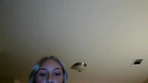 Media: Video of a blonde woman with fair skin and light makeup, looking down from above. She has a neutral expression. The background shows a ceiling with a light fixture and vent.