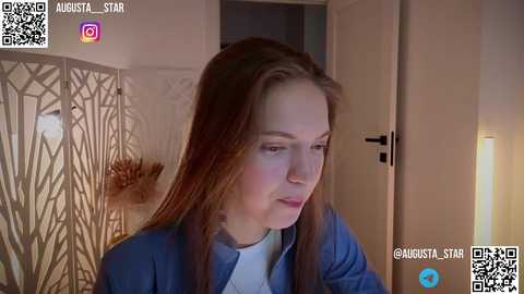 Media: A video of a young woman with long brown hair, wearing a blue jacket, sitting in a dimly lit room with a light-colored door in the background.