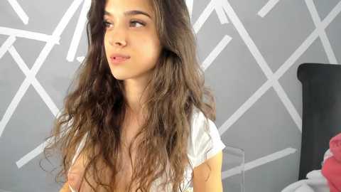 Media: Video of a young woman with long, wavy brown hair, light skin, and brown eyes, wearing a white top, gazing thoughtfully to the left. Background features geometric white patterns on a gray wall, with a hint of a black bed and red blanket in the right corner.