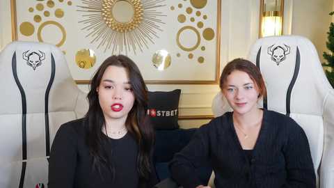 Media: Video of two young women with long dark hair, wearing black sweaters, seated in white gaming chairs with skull designs. Background features a gold sunburst mirror and stylish decor.