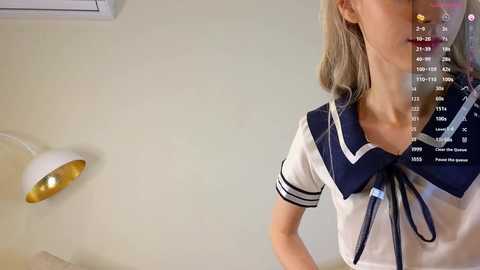 Media: Video of a blonde woman in a white sailor-style top with navy accents, standing indoors with a white lamp and beige wall in the background.