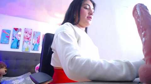 Media: Video of a woman with long black hair, wearing a white sweatshirt, playing with a large, realistic dildo in a brightly lit room with colorful anime posters on the wall.