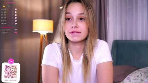 Media: Video of a young Asian girl with long blonde hair, wearing a white T-shirt, sitting in a modern bedroom with a teal headboard, a lit lamp, and a QR code in the bottom left corner.