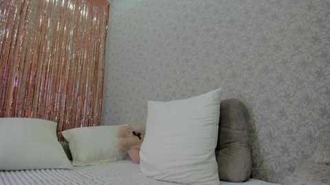 Media: Video of a woman with fair skin and blonde hair, lying on a bed with white pillows and a gray blanket. The room has a silver textured wallpaper and a shiny metallic pink curtain in the background.