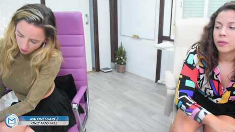 Media: A video showing two women, one in a mustard top, the other in a colorful blouse, seated in a modern office with purple chairs and light wood flooring.