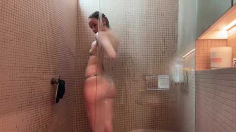 Media: A video of a nude woman with a curvy physique, fair skin, and wet hair, showering in a beige-tiled bathroom with a glass door, holding a phone to her ear.