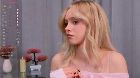 Media: Video of a young blonde woman with fair skin, wearing a pink off-shoulder top, gazing thoughtfully. Background features a gray curtain and a potted plant with pink flowers.