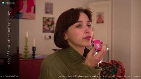 Media: Video of a fair-skinned woman with shoulder-length brown hair, wearing a green sweater, holding a pink vibrator. The background features a cozy, cluttered room with framed photos, candles, and a colorful woven bag.